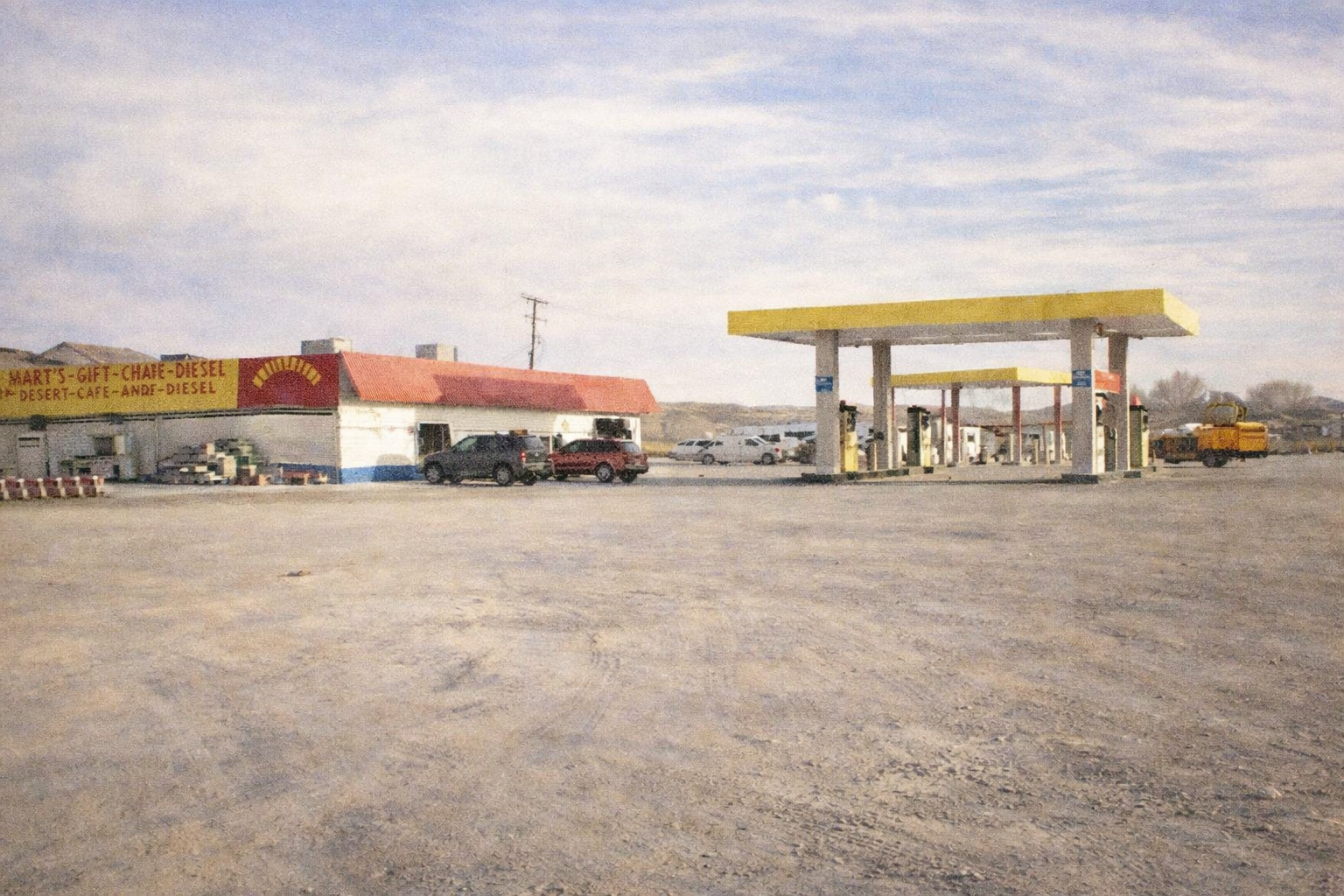 Desert gas station with yellow fuel canopies and a low building, parked vehicles nearby, set against open land and distant hills.