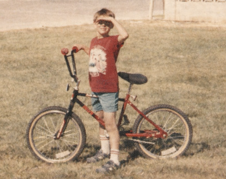 Young boy standing on grass with a red bicycle, shielding his eyes from sunlight, wearing shorts and a T-shirt in a quiet neighborhood.